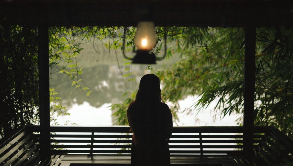Silhouette of a woman on a deck under a light looking out toward lake. She's trying to stop feeling behind.